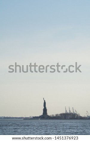 Seascape view of the Statue of Liberty silhouette in the horizon, with cranes next to it, sea in first plan and wide blue sky in background, Staten Island, Manhattan, New York, America, USA.