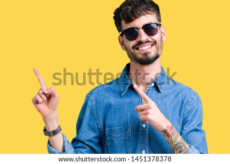 Young handsome man wearing sunglasses over isolated background smiling and looking at the camera pointing with two hands and fingers to the side.