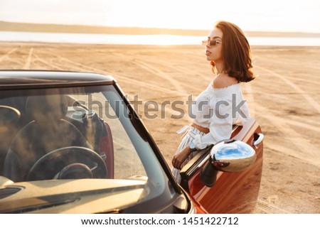 Picture of a serious young pretty woman standing near car at the beach looking aside.