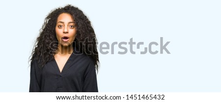 Young beautiful girl with curly hair wearing elegant dress afraid and shocked with surprise expression, fear and excited face.