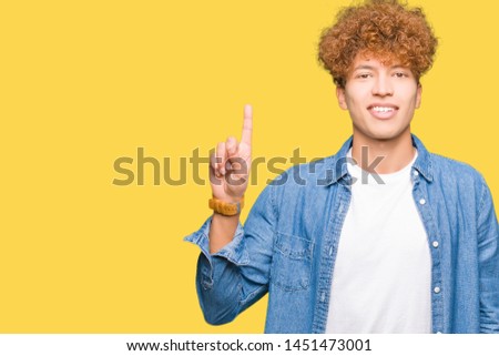 Young handsome man with afro hair wearing denim jacket showing and pointing up with finger number one while smiling confident and happy.