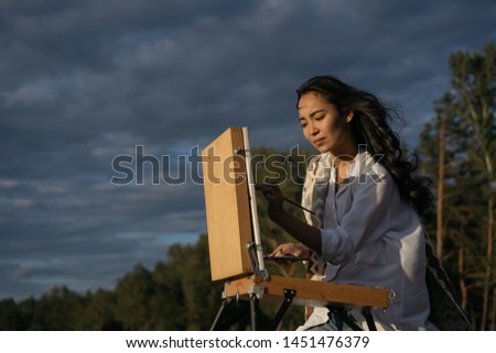 Asian woman artist painting picture on canvas outdoor. Portrait of cheerful Korean painter holding paintbrush and palette, admire beautiful landscapes and amazing sunset, looking for inspiration   