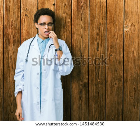 Young african american doctor woman wearing medical coat over isolated background looking stressed and nervous with hands on mouth biting nails. Anxiety problem.