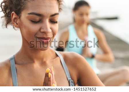 Photo of calm multiethnic sportswomen in tracksuits meditating and keeping palms together while sitting on yoga mats by seaside in morning