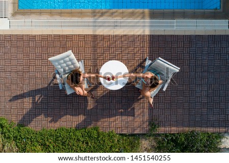 Top aerial view of women in swimsuits sunbathing by swimming pool and toasting with cocktails