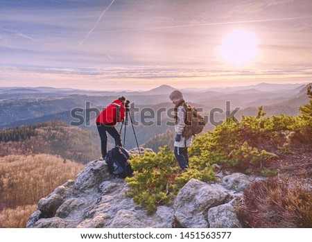 Hiker and photographer stay with tripod on cliff and takes photos. Two people stay at  tripod on the background of a mountain range and sky with clouds 