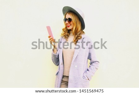 Happy smiling young woman holding phone with curly hair in round hat, pink coat jacket on wall background