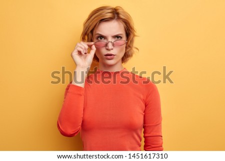 Beautiful attractive woman dressed casually looking over sunglasses at camera isolated on yellow background. close up portrait , cute girl doesn't believe in shocking news
