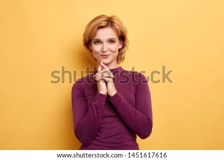 close up portrait of attractive blonde woman keeps hands together under chest as if making up plan in mind, smiling mysteriously. close up portrait. isolated yellow background