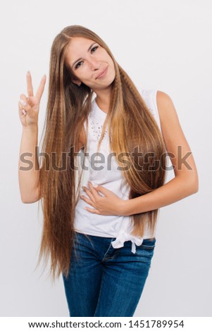 Portrait of a young stylish girl with long hair smiling, holding hands on face, is showing thumbs up victory, looking at camera. Copy space. Isolated on white background.