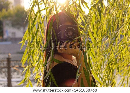 Asian Girl with camera in the park in sunset time. Brunette girl with film camera in the vintage colored image in front of willow branches