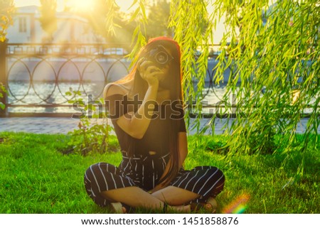 Asian Girl with camera sitting on grass cross-legged in the park in sunset time. Brunette girl with film camera in the vintage colored image