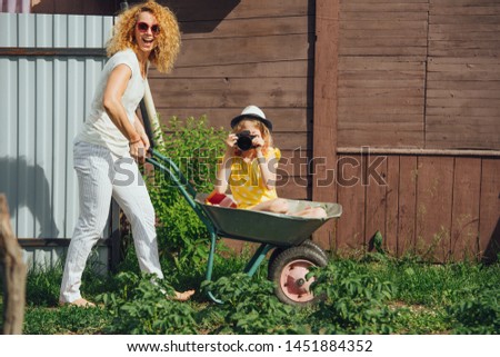 Happy mother driving a wheelbarrow, while her teenage daughter riding in it, looking at her camera, taking picture. Summer recreation in the gardens next to a country house.