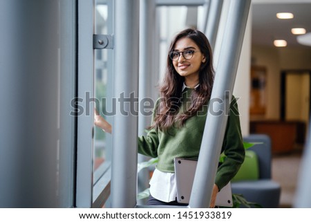 Portrait of a beautiful, young, elegant and attractive Indian Asian woman student in a preppy green sweater and spectacles. She is relaxing in a glass building during the day. 