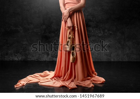 Ballerina. A young  graceful dancer standing against a black wall  dressed in a long peach dress, hands down holding pointe shoes with ribbons. Ballet studio. 
