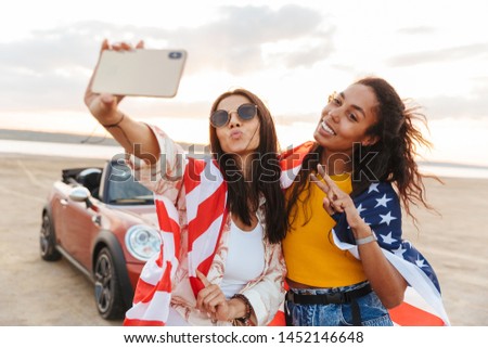 Picture of amazing young happy smiling women friends posing near car outdoors at the beach take a selfie by mobile phone holding USA flag.