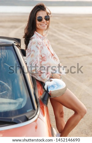 Picture of a cheerful optimistic smiling young pretty woman standing near car at the beach.