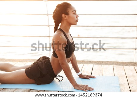 Photo of slim woman wearing sportive clothes stretching her body on finess mat while doing workout by seaside in morning Royalty-Free Stock Photo #1452189374
