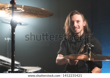 Portrait of a smiling long-haired drummer with chopsticks in his hands sitting behind a drum set. Low key. Concepts of the creative freedom of the millenial generation
