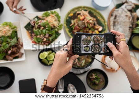 Close-up of female hands in Thailand food restaurants, shooting with mobile phones. Sea papaya salad, Grilled fish, Spicy clam leaning  are placed on white wooden table, Food photography by Smartphone