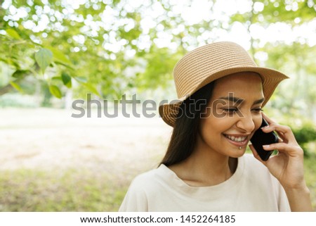 Photo of happy woman wearing straw hat and lip piercing talking on smartphone in green park