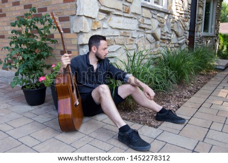 Horizontal full length view of handsome clean cut young man sitting next to home relaxing during summer morning holding acoustic guitar