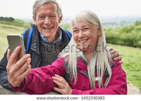 Smiling Senior Couple Hiking In Countryside Standing By Gate  And Taking Selfie On Mobile Phone     