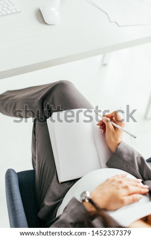 cropped view pregnant woman taking notes in notebook and holding belly while sitting in office in office chair 