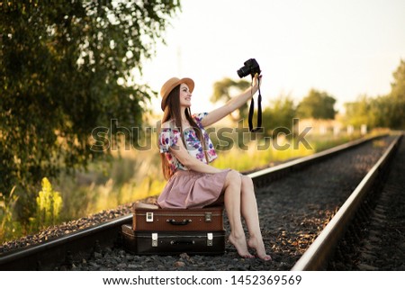 a slender girl in a straw hat is sitting on a suitcases on the railroad tracks, holding a camera and taking pictures of herself. The concept of travel, happiness, freedom, youth. Summer evening, sunse