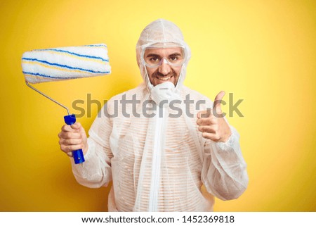 Young man wearing painter equipment and holding painting roller over isolated yellow background happy with big smile doing ok sign, thumb up with fingers, excellent sign
