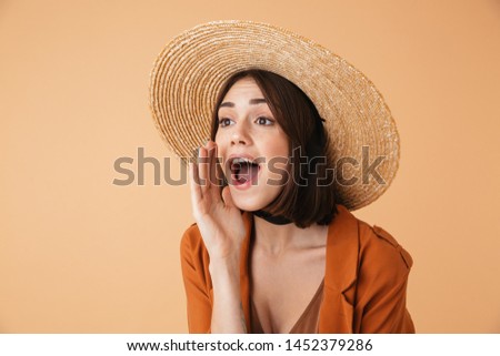 Beautiful young woman wearing straw hat and summer outfit standing isolated over beige background, shouting loud