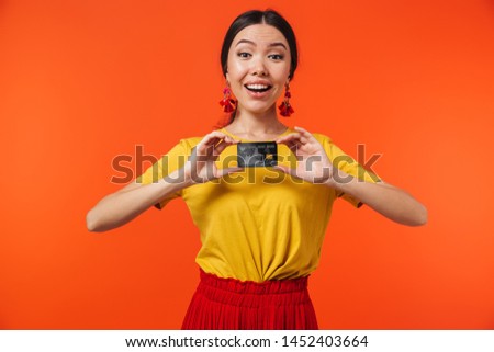 Image of surprised hispanic woman 20s dressed in skirt smiling and holding credit card isolated over red background
