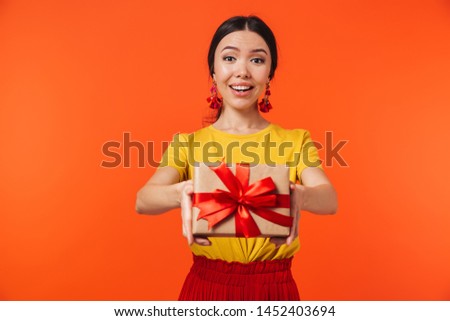 Image of joyful hispanic woman 20s dressed in skirt smiling and holding birthday present with bow isolated over red background