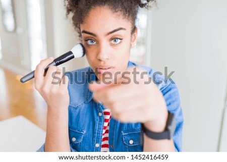 Young african american girl appying blush using make up brush pointing with finger to the camera and to you, hand sign, positive and confident gesture from the front