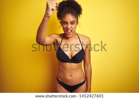African american woman on vacation wearing bikini standing over isolated yellow background angry and mad raising fist frustrated and furious while shouting with anger. Rage and aggressive concept.