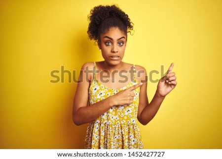 African american woman wearing casual floral dress standing over isolated yellow background Pointing aside worried and nervous with both hands, concerned and surprised expression