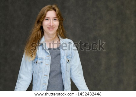 Waist up portrait of a young pretty brunette girl woman with beautiful long hair on a gray background in a jeans jacket. He talks, smiles, shows his hands with emotions in various poses.