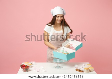 Housewife female chef cook confectioner or baker in apron white t-shirt, toque chefs hat packaging cake cupcake at table isolated on pink pastel background in studio. Mock up copy space food concept