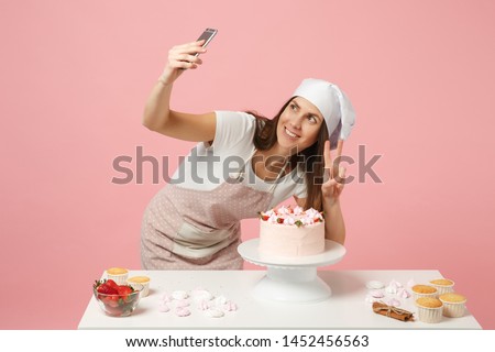 Chef cook confectioner or baker in apron white t-shirt, toque chefs hat cooking cake at table doing selfie on mobile phone isolated on pink pastel background in studio. Mock up copy space concept