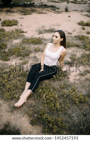 beautiful brunette girl sitting on the dry ground with grass on the background of old military aircraft. Girl in a white T-shirt and black pants barefoot in nature. Military equipment.
