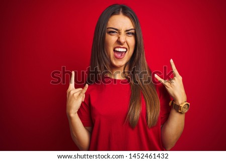Young beautiful woman wearing t-shirt standing over isolated red background shouting with crazy expression doing rock symbol with hands up. Music star. Heavy concept.