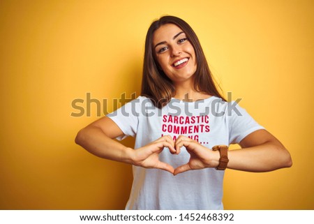 Beautiful woman wearing fanny t-shirt with irony comments over isolated yellow background smiling in love showing heart symbol and shape with hands. Romantic concept.