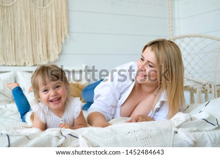 The little girl is resting and having fun on the bed in the bedroom with her mother. Bright bedroom in Scandinavian style. Macrame on the wall, wicker pillows, white blanket.