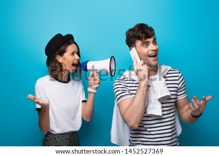 angry girl quarreling in loudspeaker at boyfriend talking on smartphone on blue background