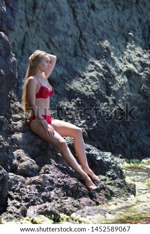 Young woman posing on coastal rock. Long-haired female person in red bikini sits on rock near waters edge 