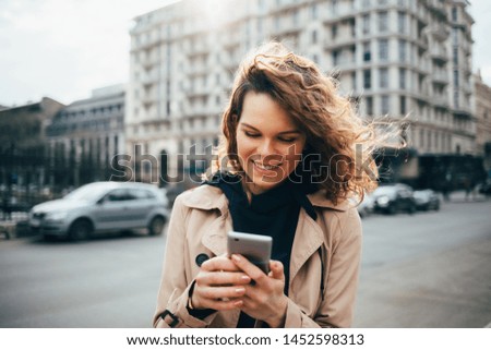 Laughing young woman wearing beige coat using smart phone standing in city near road with cars. Female typing text messages on mobile device outdoors on sunny day.
