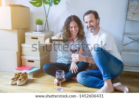 Middle age senior romantic couple in love sitting on the apartment floor with boxes around, celebrating drinking a glass of wine smiling happy for moving to a new home