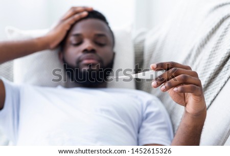 Fever, seasonal flu. Sick african-american man lying on sofa and checking his temperature with thermometer Royalty-Free Stock Photo #1452615326