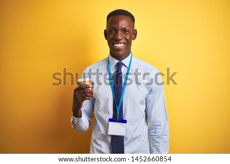 African american businessman wearing id drinking coffee over isolated yellow background with a happy face standing and smiling with a confident smile showing teeth