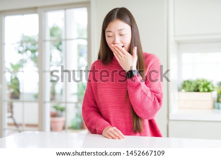 Beautiful Asian woman wearing pink sweater on white table bored yawning tired covering mouth with hand. Restless and sleepiness.